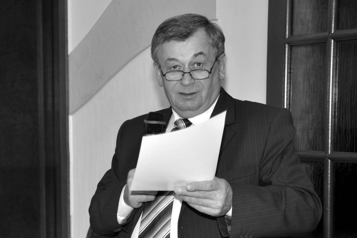 Czarno-biała fotografia przedstawia mężczyznę w garniturze i okularach, stojącego przy mikrofonie. Trzyma kartkę z przygotowanym tekstem i przemawia podczas uroczystości odbywającej się we wnętrzu budynku.